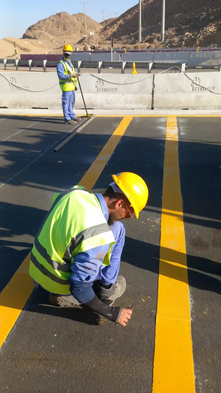 Workers applying yellow road markings on asphalt beside Petrojet barriers.